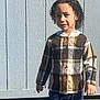 child, curly_hair, plaid_shirt, jeans, standing, outdoor, sunlight, smile, fence, wooden_fence, casual_clothing, portrait, young, person, daylight, shadow, happy, front_view, clothing, nature