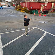 Adrian is registered to the contest to win money with this photo: autumn, boy, buildings, casual_clothing, child, confident, curly_hair, daylight, jacket, orange_cones, outdoor, parking_lot, pavement, playful, sidewalk, sneakers, stop_sign, street, toy_gun, urban