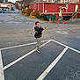 autumn, boy, buildings, casual_clothing, child, confident, curly_hair, daylight, jacket, orange_cones, outdoor, parking_lot, pavement, playful, sidewalk, sneakers, stop_sign, street, toy_gun, urban