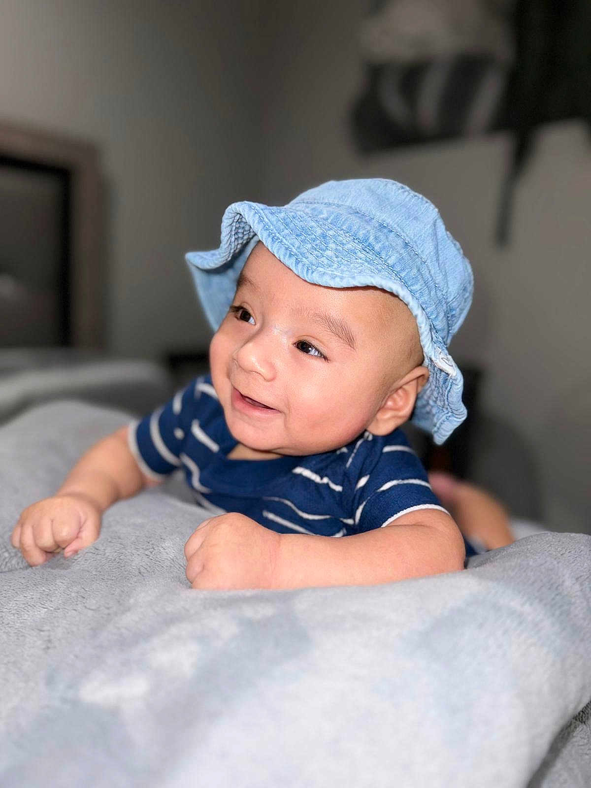 William joined the competition — help win amazing prizes! baby, child, smile, hat, denim_hat, striped_shirt, blue_shirt, blanket, indoor, face, happy, cute, infant, person, skin, portrait, lying_down, soft_texture, cozy, young