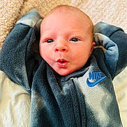 Charles is registered to the contest to win money with this photo: baby, newborn, infant, face, expression, clothing, nike, fleece, blanket, soft, cute, portrait, child, lying_down, hands_up, skin, head, eyes, mouth, indoors