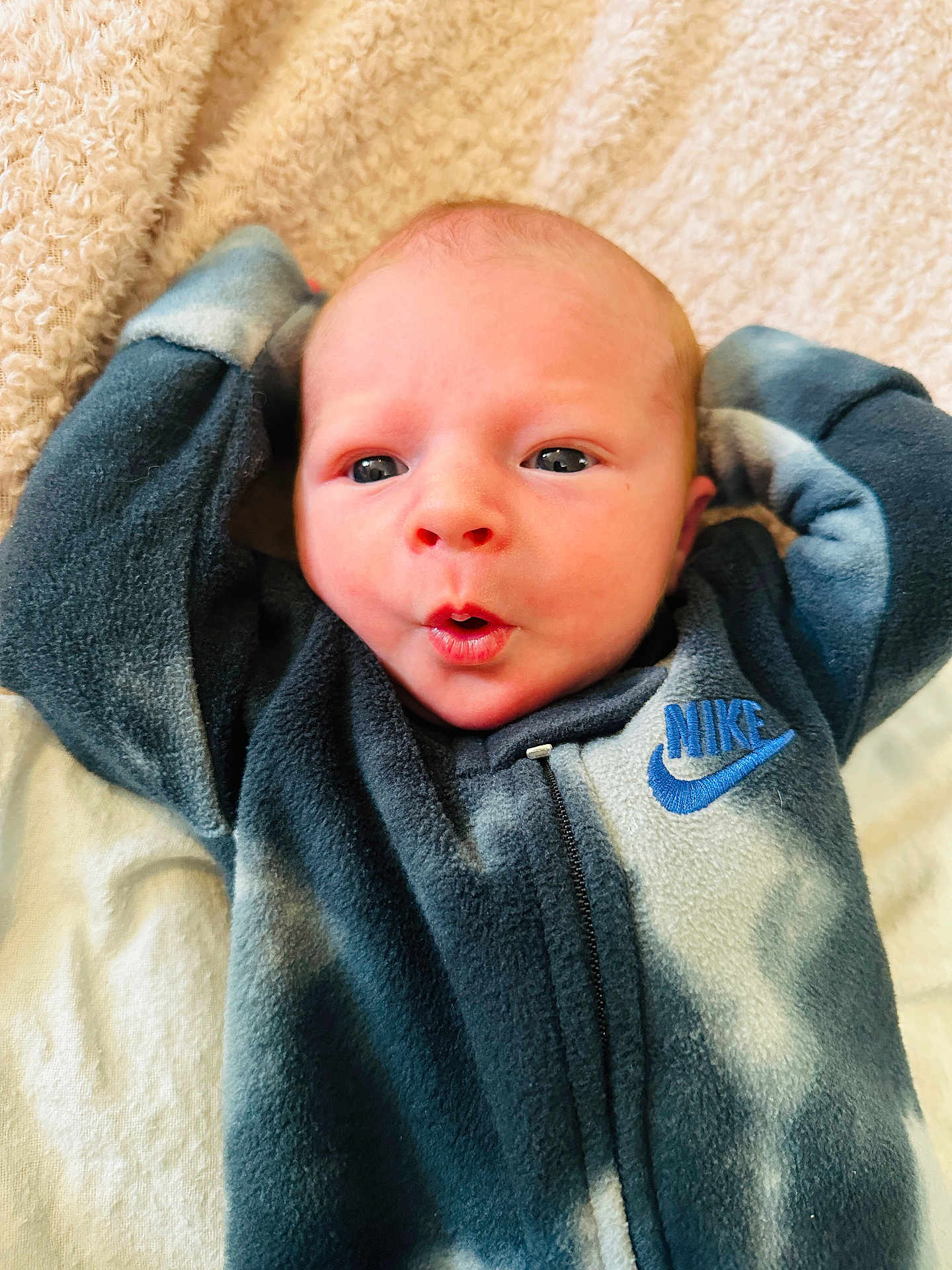 Charles is registered to the contest to win money with this photo: baby, newborn, infant, face, expression, clothing, nike, fleece, blanket, soft, cute, portrait, child, lying_down, hands_up, skin, head, eyes, mouth, indoors
