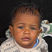 Laudrick is registered to the contest to win money with this photo: child, toddler, face, portrait, sweater, dinosaur_pattern, curly_hair, brown_skin, eyes, clothing, cute, indoors, blanket, expression, young_child, person, head, baby, sitting, casual