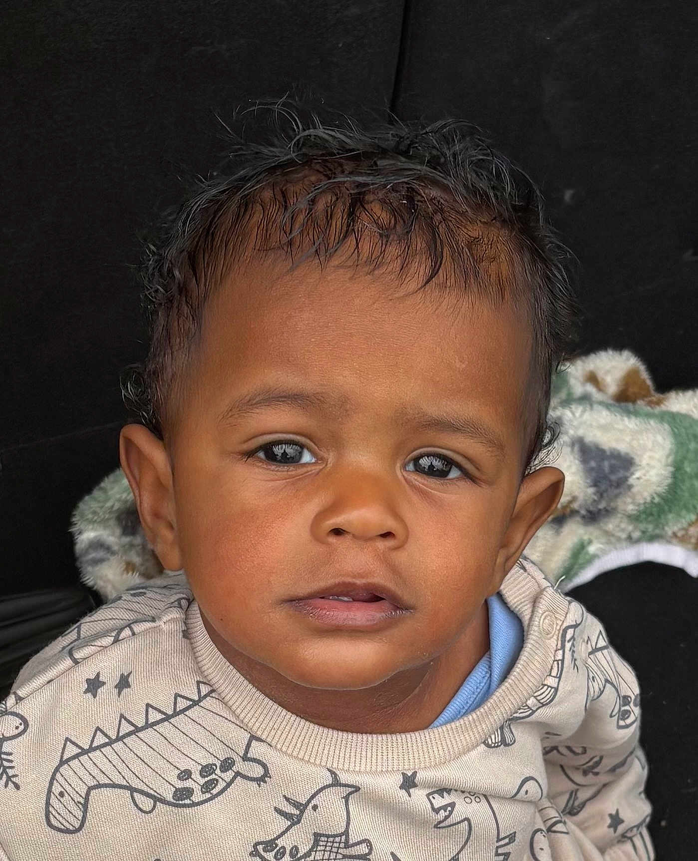 Laudrick is registered to the contest to win money with this photo: child, toddler, face, portrait, sweater, dinosaur_pattern, curly_hair, brown_skin, eyes, clothing, cute, indoors, blanket, expression, young_child, person, head, baby, sitting, casual