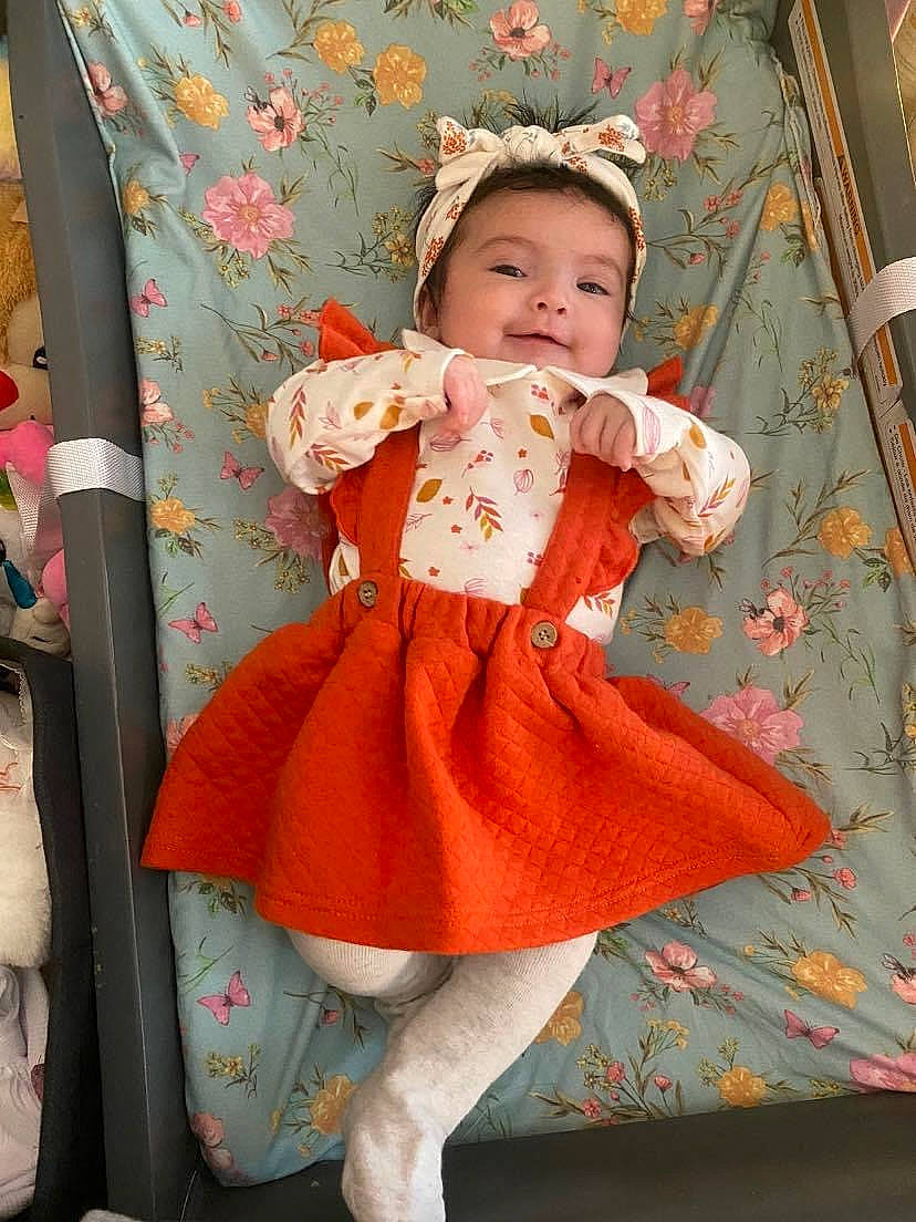 Amariah is registered to the contest to win money with this photo: baby, baby_toddler_clothing, child, day_dress, embellishment, face, happy, human_leg, linens, pattern, peach, person, pink, ruffle, sitting, sleeve, smile, sock, textile, thigh