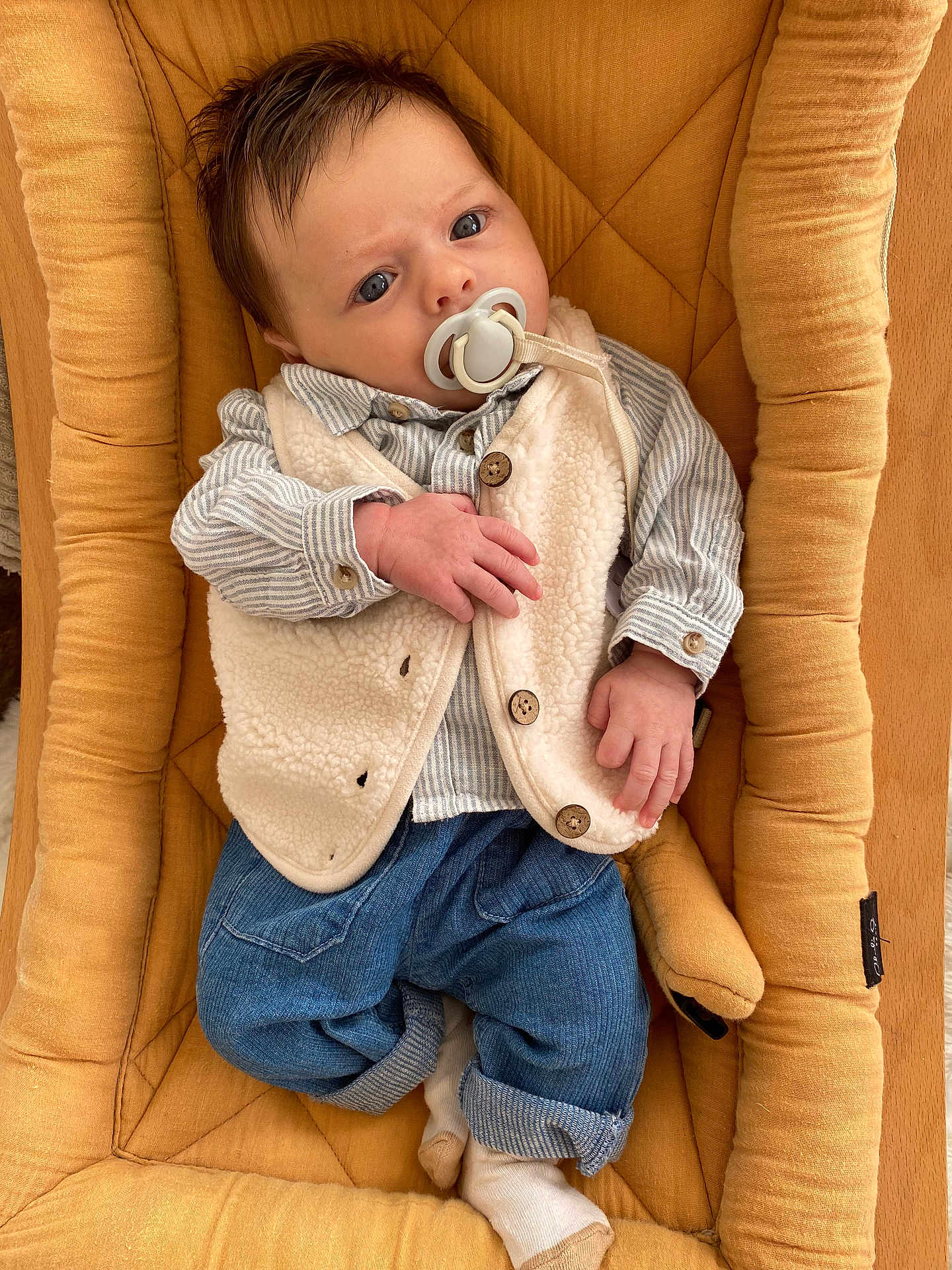 Elio a rejoint le concours — aidez-le/la à gagner de superbes lots ! baby, infant, pacifier, vest, striped_shirt, jeans, socks, cushioned_seat, mustard_color, curious_expression, indoor, young_child, clothing, portrait, relaxed, casual, hands, face, head, comfort