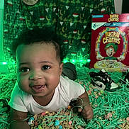 Jeremiah is registered to the contest to win money with this photo: baby, infant, cereal, lucky_charms, green_lights, festive_backdrop, shoes, smiling, lying_down, colorful, decorative_strings, indoors, cute, child, face, portrait, white_clothing, playful, closeup, happy