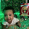 baby, infant, cereal, lucky_charms, green_lights, festive_backdrop, shoes, smiling, lying_down, colorful, decorative_strings, indoors, cute, child, face, portrait, white_clothing, playful, closeup, happy