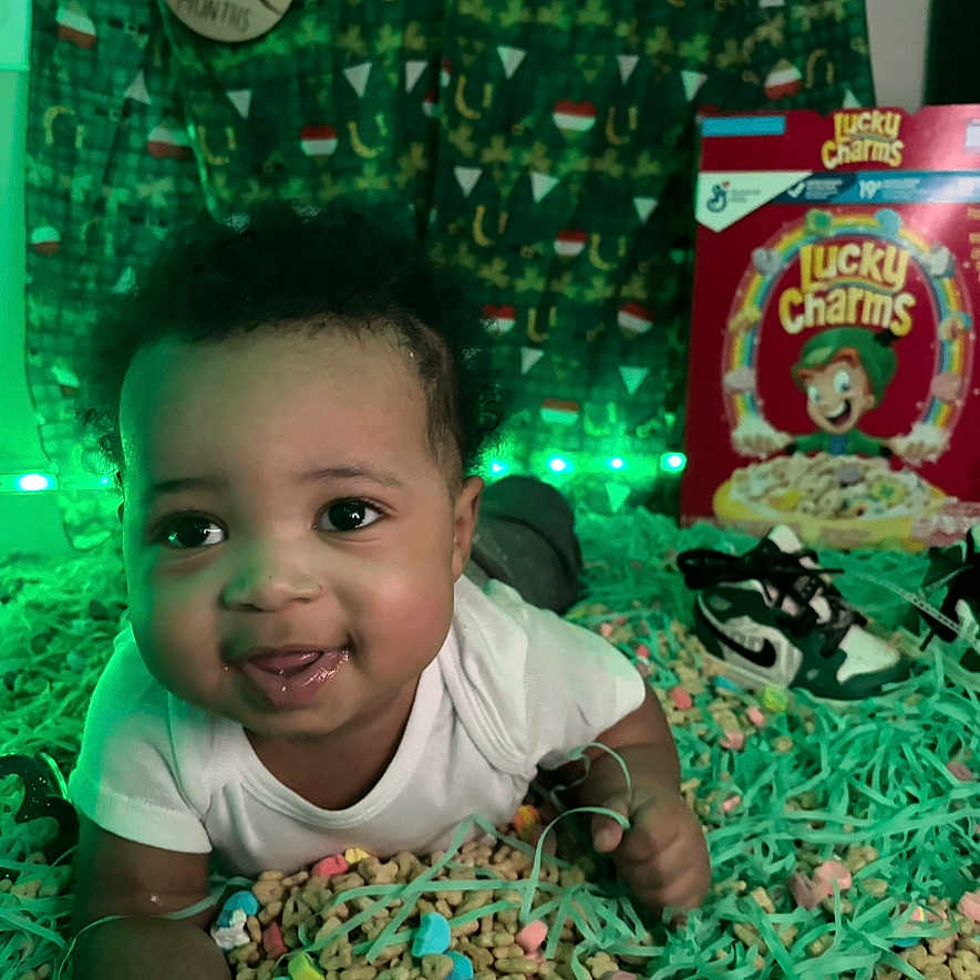 Jeremiah is registered to the contest to win money with this photo: baby, infant, cereal, lucky_charms, green_lights, festive_backdrop, shoes, smiling, lying_down, colorful, decorative_strings, indoors, cute, child, face, portrait, white_clothing, playful, closeup, happy