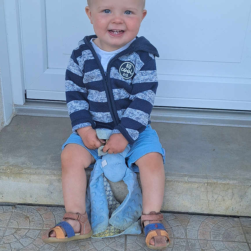 Noah a rejoint le concours — aidez-le/la à gagner de superbes lots ! blue_eyes, blue_shorts, casual_clothing, child, concrete, cute, daylight, doorstep, front_door, happy, outdoor, person, sandals, short_hair, sitting, smiling, striped_hoodie, stuffed_toy, tile_floor, toddler