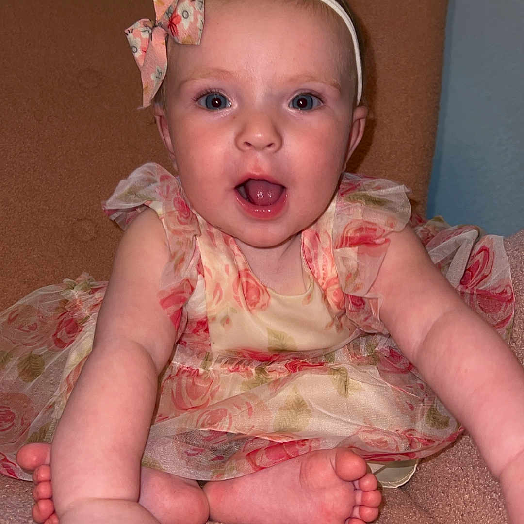 Esme is registered to the contest to win money with this photo: 6_months, baby, child, closeup, cute, feet, floral_dress, girl, hands, happy, headband, indoors, infant, person, pink_chair, portrait, reaching_out, skin, smiling, window_blinds