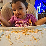 Valentina is registered to the contest to win money with this photo: toddler, child, high_chair, messy, spaghetti, food, meal_time, indoors, face, hands, smile, clothing, purple_shirt, tray, television, home, happy, candid, babysitter, family