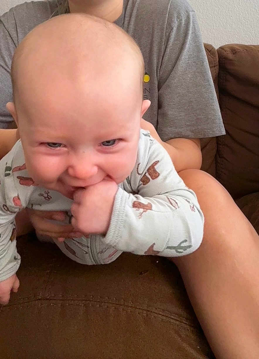 Trinity is registered to the contest to win money with this photo: baby, child, infant, hand, couch, person, clothing, onesie, animal_print, face, smile, chewing, adult, holding, arm, sitting, indoor, casual, fabric, skin