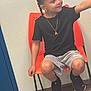 child, braided_hair, orange_chair, black_tshirt, gray_shorts, black_sneakers, gold_necklace, indoor, smile, sitting, floor, wall, person, casual_clothing, shorts, footwear, young, happy, portrait, chair