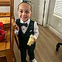 child, boy, banana, smile, suit, bow_tie, indoor, wooden_furniture, floor, door, window_blinds, white_shirt, shoes, happy, person, portrait, standing, hand, face, hair