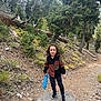 child, smiling, curly_hair, rock, forest, trees, path, outdoor, toy, shovel, nature, hiking, casual_clothing, black_pants, colorful_shirt, footwear, daytime, greenery, adventure, happy