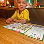 child, yellow_shirt, restaurant, booth, table, coloring, placemat, activity_sheet, crayon, smiling, indoor, wood, lighting, face, person, happy, clothing, dining, casual, kid