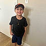 child, boy, smile, black_tshirt, shorts, baseball_cap, indoor, wooden_floor, beige_wall, necklace, gold_pendant, standing, casual_clothing, happy, portrait, young, person, feet, socks, shadow