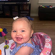 Kaia is registered to the contest to win money with this photo: baby, smiling, child, blue_eyes, hair_bow, sitting, baby_seat, butterfly_pattern, indoor, living_room, television, flooring, toy, happy, face, person, clothing, furniture, earrings, cute