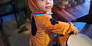 Abel is registered to the contest to win money with this photo: toddler, child, costume, dog_costume, indoor, table, wooden_table, basket, plant, floor, curious, looking_back, face, clothing, baby, furniture, home, childhood, cute, person
