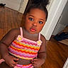 child, toddler, baby, portrait, sitting, indoors, doorway, wooden_floor, colorful_clothing, striped_outfit, pink_clothing, hair_bun, curly_hair, serious_expression, looking_at_camera, face, arms, legs, home, photography