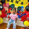 child, toddler, birthday, birthday_party, balloons, cartoon, minnie_mouse, mickey_mouse, donald_duck, party_decoration, pink_shirt, jeans, pink_shoes, sitting, portrait, smiling, indoors, colorful, wooden_floor, gift_wrap