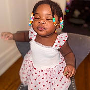 Leyana is registered to the contest to win money with this photo: child, toddler, baby, portrait, sitting, smiling, eyes_closed, pursed_lips, dress, heart_pattern, colorful_beads, braided_hair, indoor, chair, cushion, wooden_floor, home, snack, close_up, happy