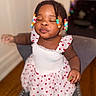 child, toddler, baby, portrait, sitting, smiling, eyes_closed, pursed_lips, dress, heart_pattern, colorful_beads, braided_hair, indoor, chair, cushion, wooden_floor, home, snack, close_up, happy