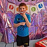 Tanner is registered to the contest to win money with this photo: child, smiling, portrait, standing, indoors, party, birthday, colorful, foil_backdrop, decorations, banners, letters, game_table, blue_shirt, black_shorts, red_hair, playful, cartoon_smile, festive, photography