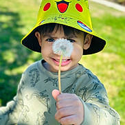 Kal is registered to the contest to win money with this photo: child, boy, hat, pikachu, yellow, dandelion, flower, hand, smile, outdoor, nature, grass, green, sunlight, portrait, casual_clothing, young, cute, playful, happy