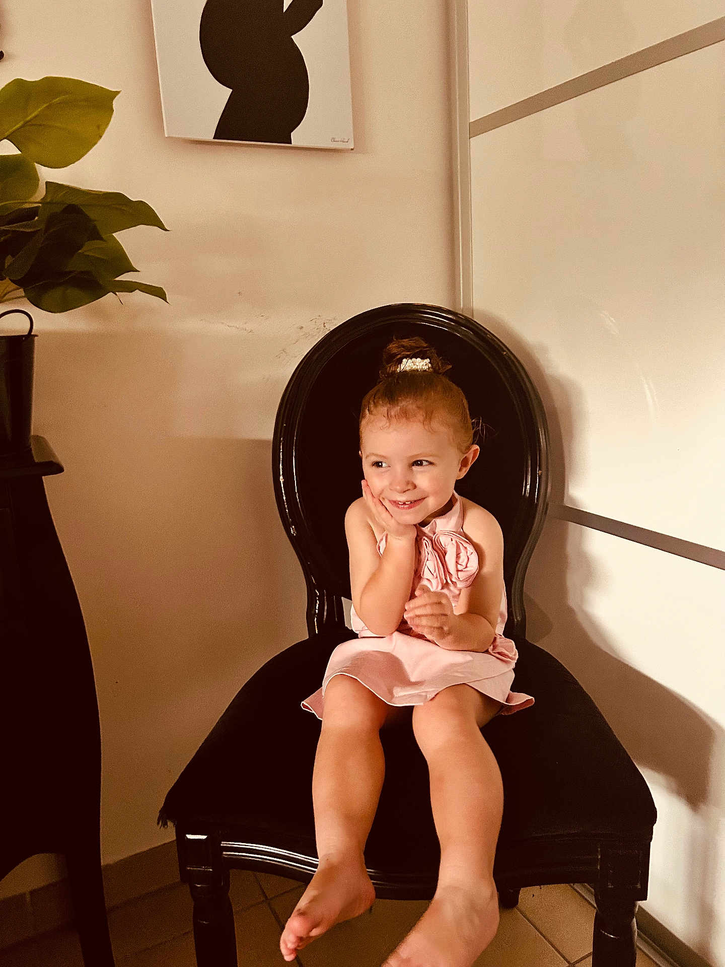 Milie participe au concours pour gagner de l'argent avec cette photo : child, girl, pink_dress, barefoot, black_chair, smile, indoor, plant, artwork, floor_tiles, cute, happy, portrait, seated, young_child, decor, furniture, bare_legs, hair_bun, light_shadow