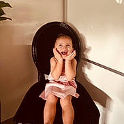 Milie participe au concours pour gagner de l'argent avec cette photo : toddler, child, pink_dress, black_chair, smiling, happy, indoor, furniture, plant, portrait, cute, young_child, seated, hands_on_face, joyful, expression, home, wall, light_shadow, casual