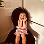 toddler, child, pink_dress, black_chair, smiling, happy, indoor, furniture, plant, portrait, cute, young_child, seated, hands_on_face, joyful, expression, home, wall, light_shadow, casual