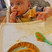 Kayden a rejoint le concours — aidez-le/la à gagner de superbes lots ! baby, high_chair, baby_food, messy, orange_food, face, hand, spoon, tray, feeding, child, blue_eyes, clothing, curtain, window, indoor, infant, cute, expression, person