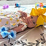 baby, infant, yellow_bow, headband, lemon_pattern, dress, blue_flowers, blanket, toy, lying_down, cute, smiling, face, skin, child, soft_focus, indoor, decorations, colorful, happy