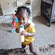 Junie is registered to the contest to win money with this photo: toddler, child, baby, drinking, bottle, yellow_outfit, curly_hair, smiling, portrait, indoors, living_room, carpet, rug, toy, playpen, furniture, cabinet, standing, home, candid