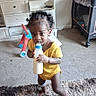 toddler, child, baby, drinking, bottle, yellow_outfit, curly_hair, smiling, portrait, indoors, living_room, carpet, rug, toy, playpen, furniture, cabinet, standing, home, candid