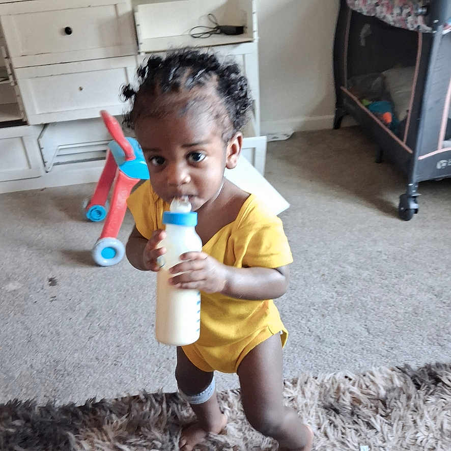 Junie is registered to the contest to win money with this photo: toddler, child, baby, drinking, bottle, yellow_outfit, curly_hair, smiling, portrait, indoors, living_room, carpet, rug, toy, playpen, furniture, cabinet, standing, home, candid