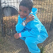 J'Hleel joined the competition — help win amazing prizes! child, smiling, outdoor, animal_enclosure, fence, blue_tracksuit, sneakers, crouching, portrait, boy, short_hair, jewelry, watch, bracelet, straw, dirt, playground, tunnel, daylight, happy