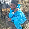 child, smiling, outdoor, animal_enclosure, fence, blue_tracksuit, sneakers, crouching, portrait, boy, short_hair, jewelry, watch, bracelet, straw, dirt, playground, tunnel, daylight, happy