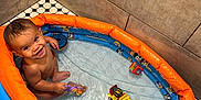 Jaden is registered to the contest to win money with this photo: bathroom, blue, child, corner, cute, fun, happy, indoor, inflatable_pool, orange, play, purple_toy, sitting, smiling, tiles, toddler, toy, water, wet, yellow_truck