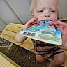 Everett is registered to the contest to win money with this photo: baby, barcode, barefoot, blond_hair, blue_eyes, child, close_up, colorful_packaging, curious, face, gravel, hands, indoors, package, portrait, sitting, striped_clothing, toddler, wide_angle, wooden_structure