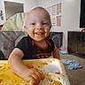 Everett is registered to the contest to win money with this photo: baby, black_shirt, child, dining, face, food, hands, happy, high_chair, indoor, living_room, meal, messy, orange_food, playroom, portrait, seated, smiling, television, toddler