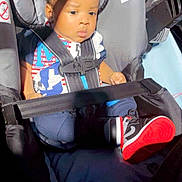Elliott is registered to the contest to win money with this photo: baby, car_seat, child, infant, seatbelt, clothing, sneakers, footwear, curly_hair, sunlight, shadow, sitting, indoor, safety, cute, person, young_child, black_hair, casual_wear, portrait