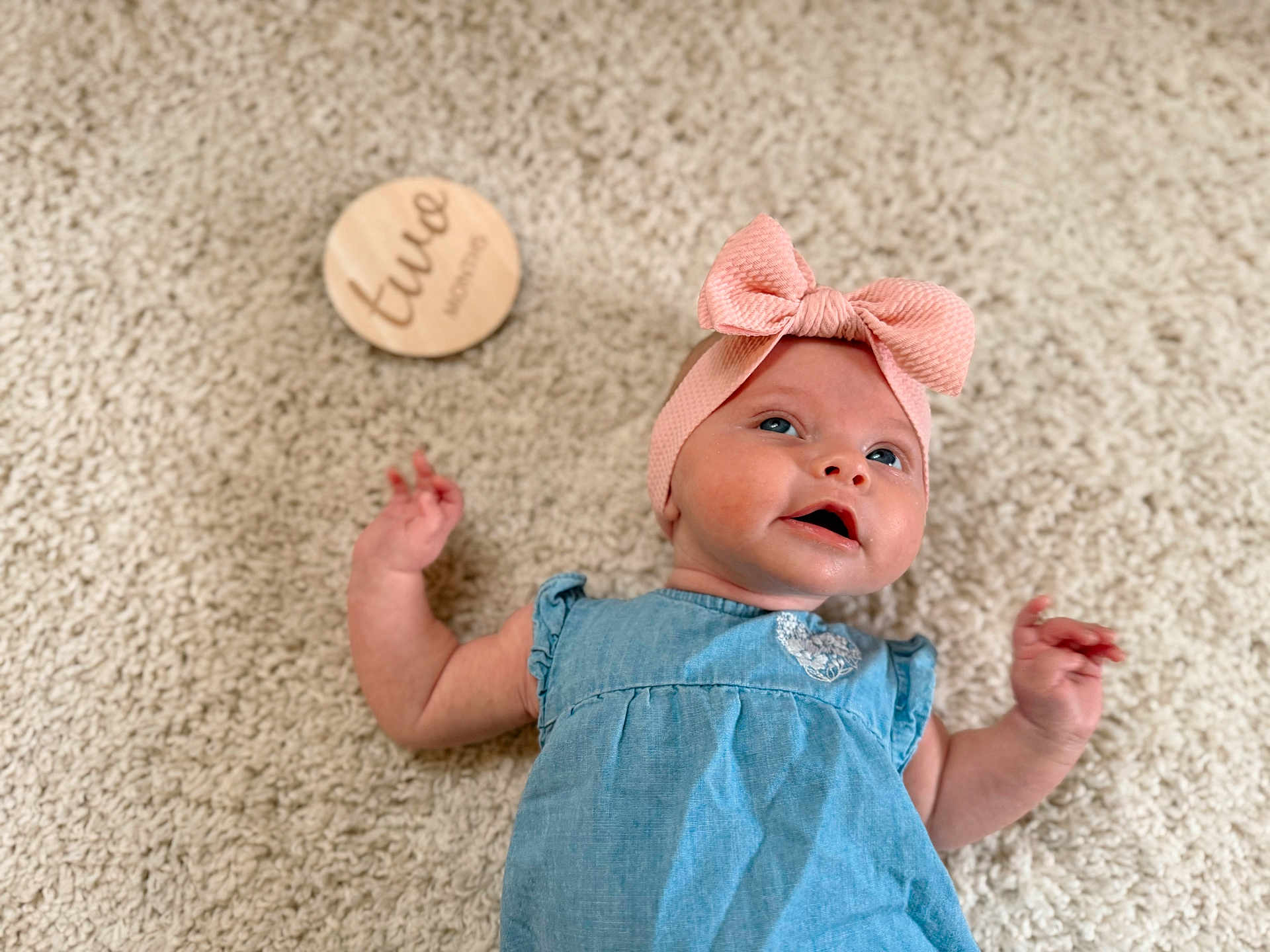 Olenna is registered to the contest to win money with this photo: baby, child, infant, blue_dress, pink_bow, headband, carpet, floor, wooden_circle, milestone, cute, portrait, smiling, happy, indoor, skin, hand, face, lying_down, closeup