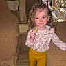 child, girl, indoor, curly_hair, bow, snack, yellow_pants, pink_shoes, patterned_top, furniture, wooden_floor, cozy, smiling, standing, home, curtains, table, chair, person, portrait