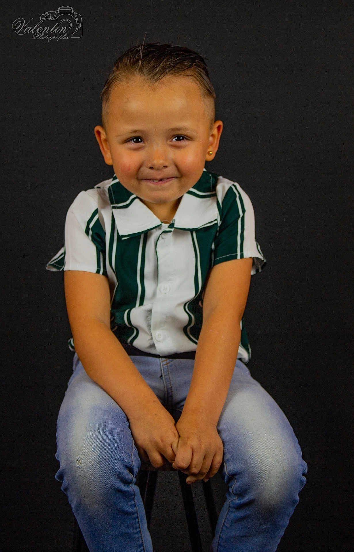 Wesley a rejoint le concours — aidez-le/la à gagner de superbes lots ! child, boy, portrait, smiling, studio, dark_background, striped_shirt, green_shirt, jeans, sitting, stool, short_hair, earring, hands_clasped, cute, playful_expression, one_person, photography, watermark, indoor