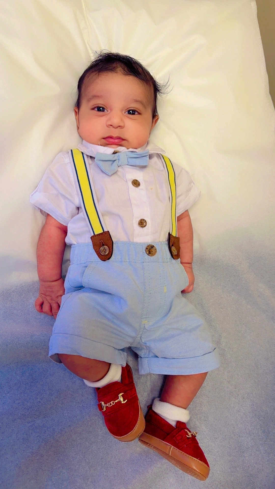 Avik is registered to the contest to win money with this photo: baby, infant, bow_tie, suspenders, red_shoes, socks, shorts, shirt, buttons, lying_down, portrait, cute, eyes_open, hands, feet, blanket, soft_lighting, white_background, fashion, outfit
