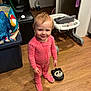 toddler, child, pink_onesie, polka_dot, smile, standing, wooden_floor, high_chair, toy, storage_bin, household, indoor, floor, person, face, footwear, curly_hair, baby, room, playful