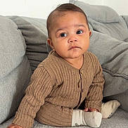 Anastasya a rejoint le concours — aidez-le/la à gagner de superbes lots ! baby, child, infant, sitting, brown_clothing, ribbed_sweater, grey_blanket, grey_cushions, indoor, portrait, cute, young_child, cozy, soft_texture, hand, face, expression, looking_away, sock, floor